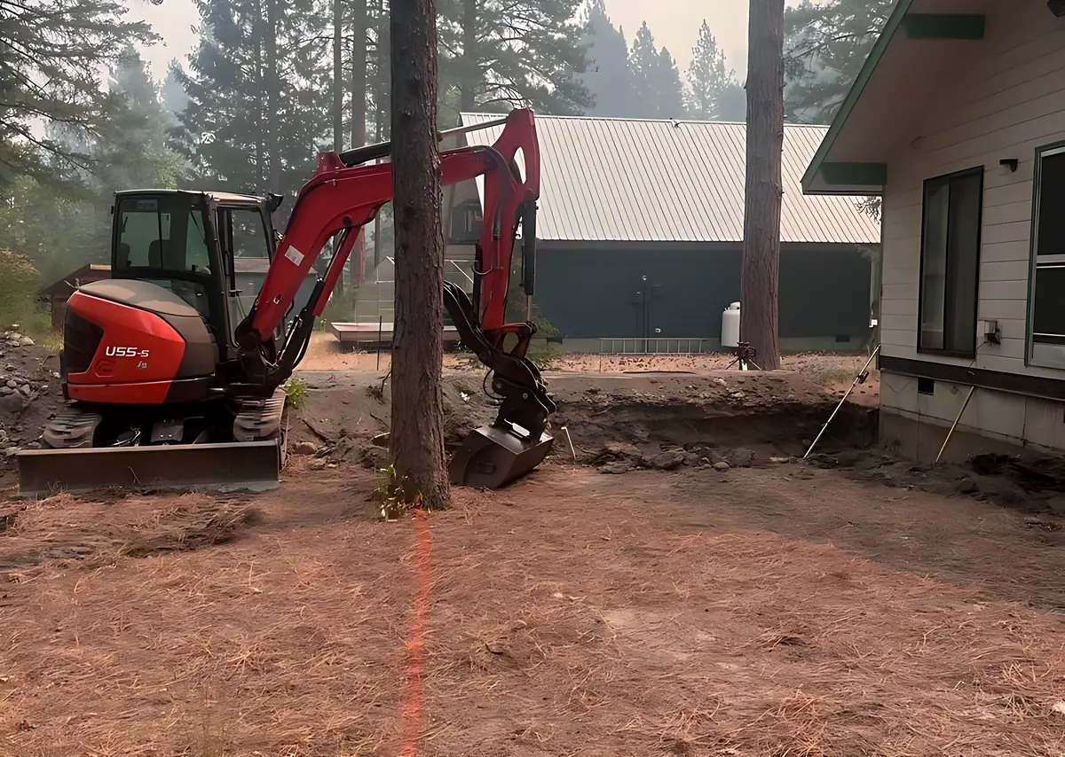 Kubota U55-5 clearing ground between pines beside a house, with a marking line painted on the ground