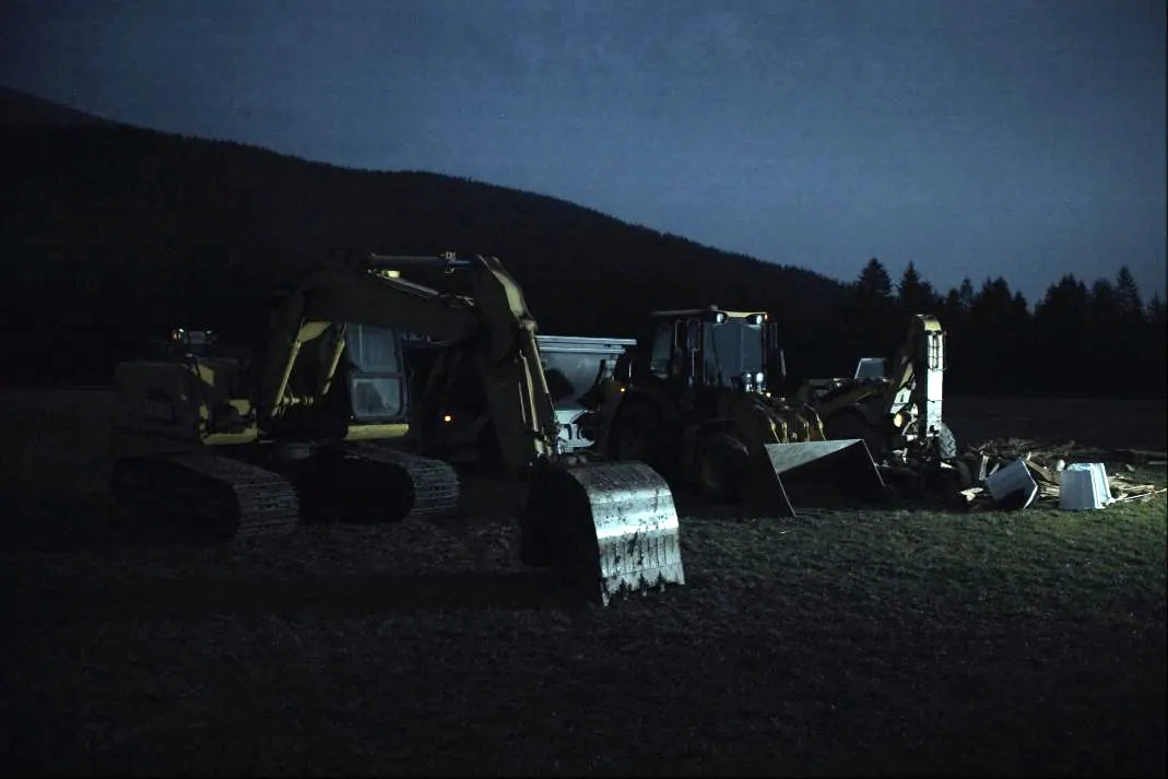 Excavator, compact track loader and trailer parked at the edge of a jobsite at dusk.
