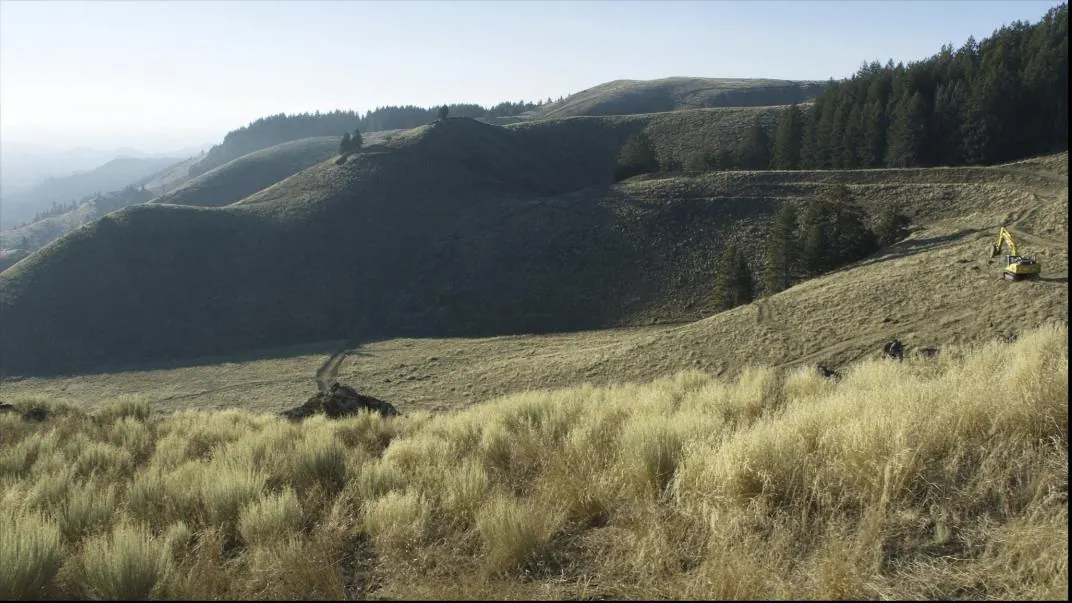 Raw sagebrush and pine hillside with a single excavator beginning the first cut.