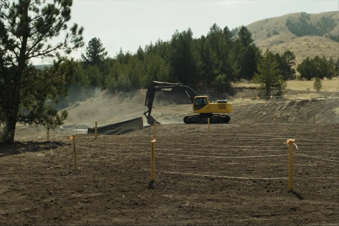 An excavator stripping topsoil and shaping a level building pad, with grade stakes set.