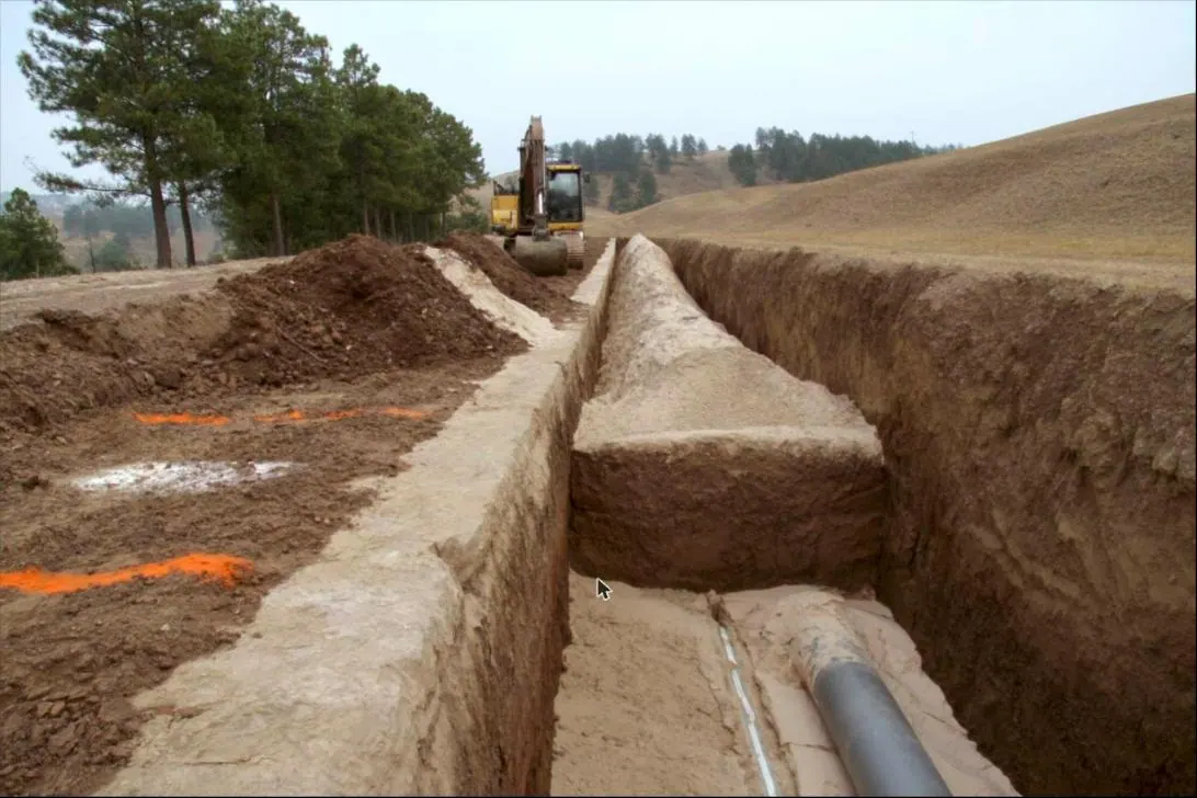 An open utility trench with conduit and a water line bedded in clean sand, spoil windrowed alongside.