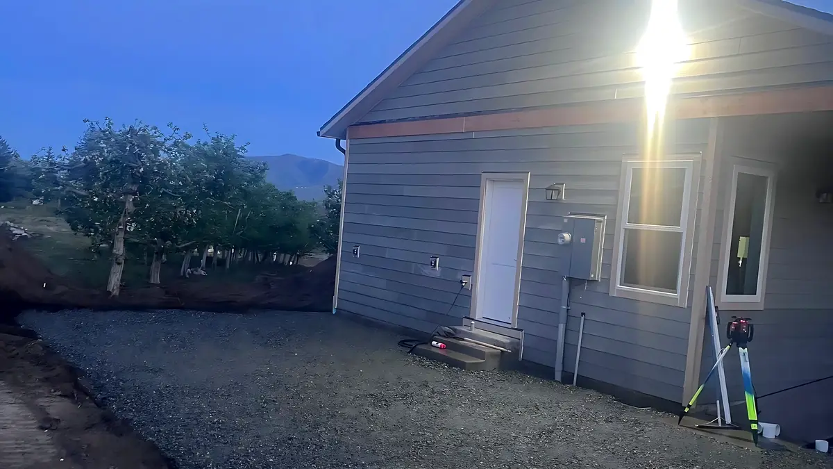 Graded crushed rock pad along a new house at dusk with a laser level on its tripod